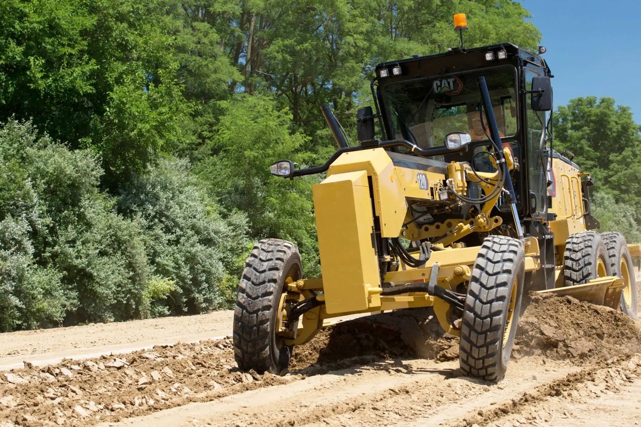 Yellow motor grader operating at a construction site surrounded by greenery in India.